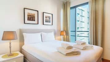 Habitación de hotel con una cama, dos cuadros en la pared, una mesita de noche con lámpara y una ventana con cortinas.
