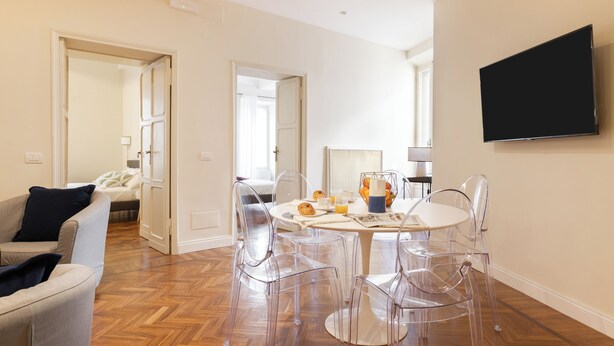 A modern living room with a wooden herringbone floor, a round dining table with clear chairs, a flat-screen TV mounted on the wall, and a sofa with a dark cushion. - Coppelle Charme - Three Bedroom Apartment, Sleeps 6 (Rome)