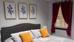 A neatly made bed with a patterned quilt, two pillows, and a rolled towel on a dark headboard. Three framed floral artworks are hung on the wall above the bed. - A beautiful 4 BDR home, newly renovated and close to everything! (Williston)