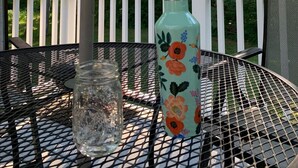 A metal table with a glass jar and a floral water bottle under a green umbrella, set on a deck with a white railing and a forest backdrop. - A beautiful 4 BDR home, newly renovated and close to everything! (Williston)