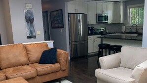 A modern living room with a brown sofa and a white armchair, a kitchen with stainless steel appliances, and a large window. - A beautiful 4 BDR home, newly renovated and close to everything! (Williston)