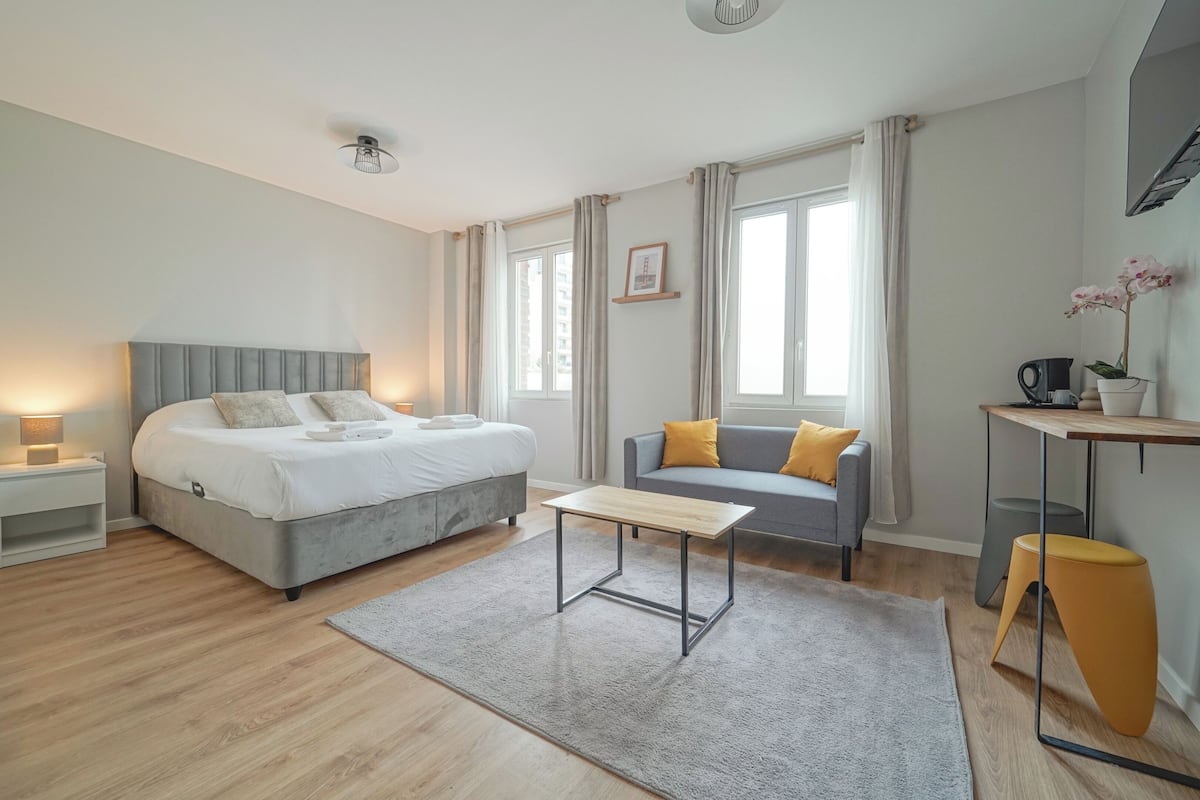 Egy modern hálószoba, amelyben található egy ágy, egy éjjeliszemélyzet, egy kis íróasztal, egy kanapé és egy ablak, amelyen függöny van.