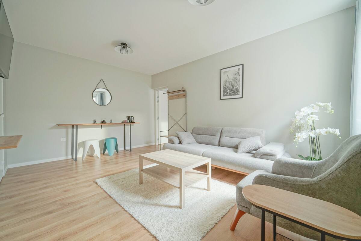Egy modern nappali, amelyben van egy kanapé, egy dohányzóasztal és egy kisebb, mellékes asztal. Az egyik falon tükör, a kanapé felett pedig egy kép található keretben.
