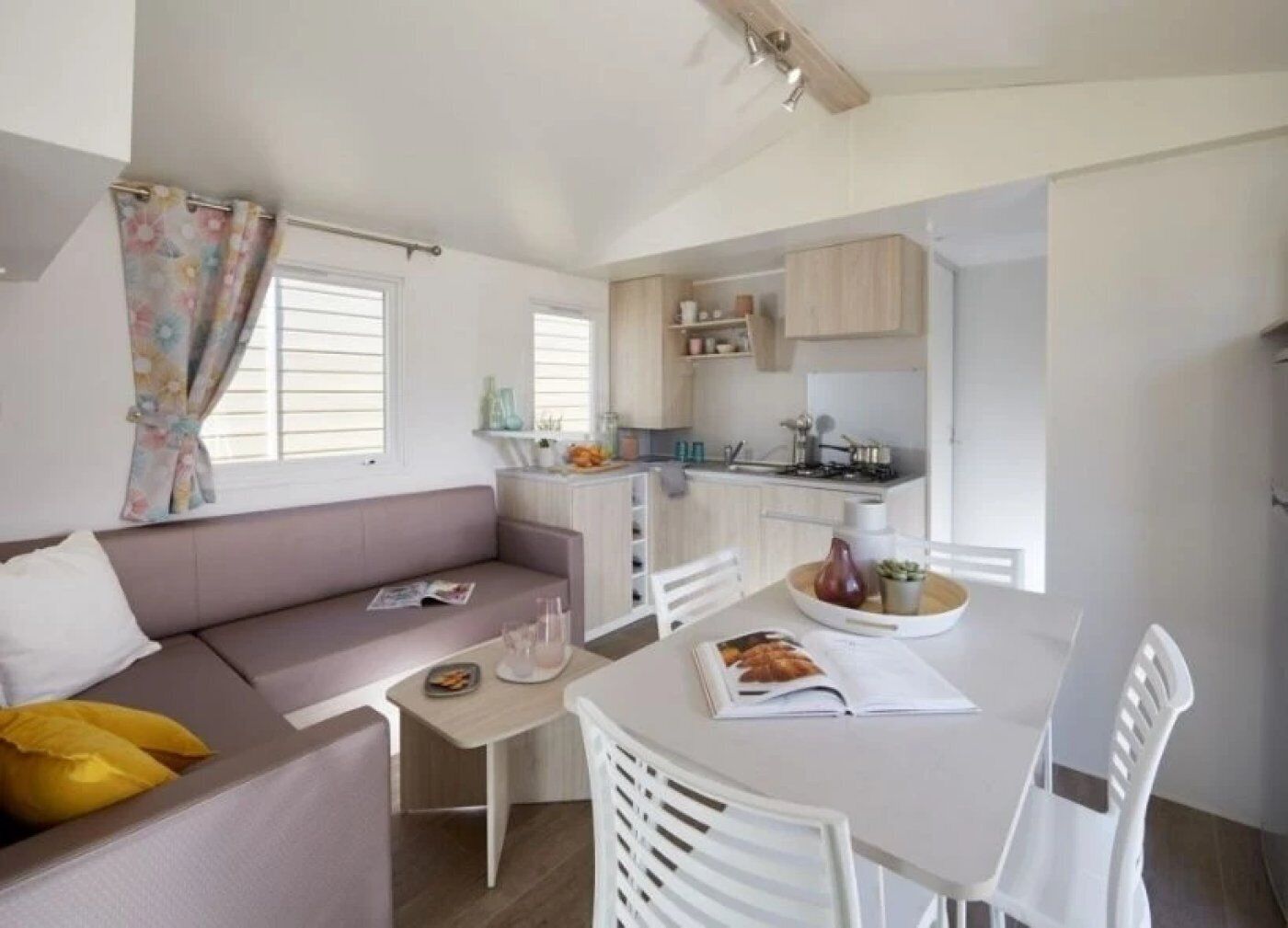 Un espacio habitable compacto con sofá, mesa de comedor, sillas, una pequeña cocina y una ventana con cortinas.