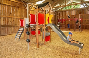 A wooden play structure with slides and climbing elements, surrounded by a covered area with a wooden floor.