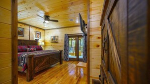 Interior de una cabaña de madera con una cama, un ventilador de techo y un televisor.