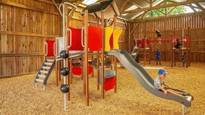 A wooden play structure with slides and climbing elements, surrounded by a covered area with a wooden floor.