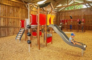 Une structure de jeux en bois, dotée de toboggans et d’éléments d’escalade, entourée d’une zone abritée avec un plancher en bois.