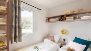 Ein Schlafzimmer mit zwei Betten, einem Fenster mit Vorhängen, einem Regal mit Büchern und einem kleinen runden Tisch.