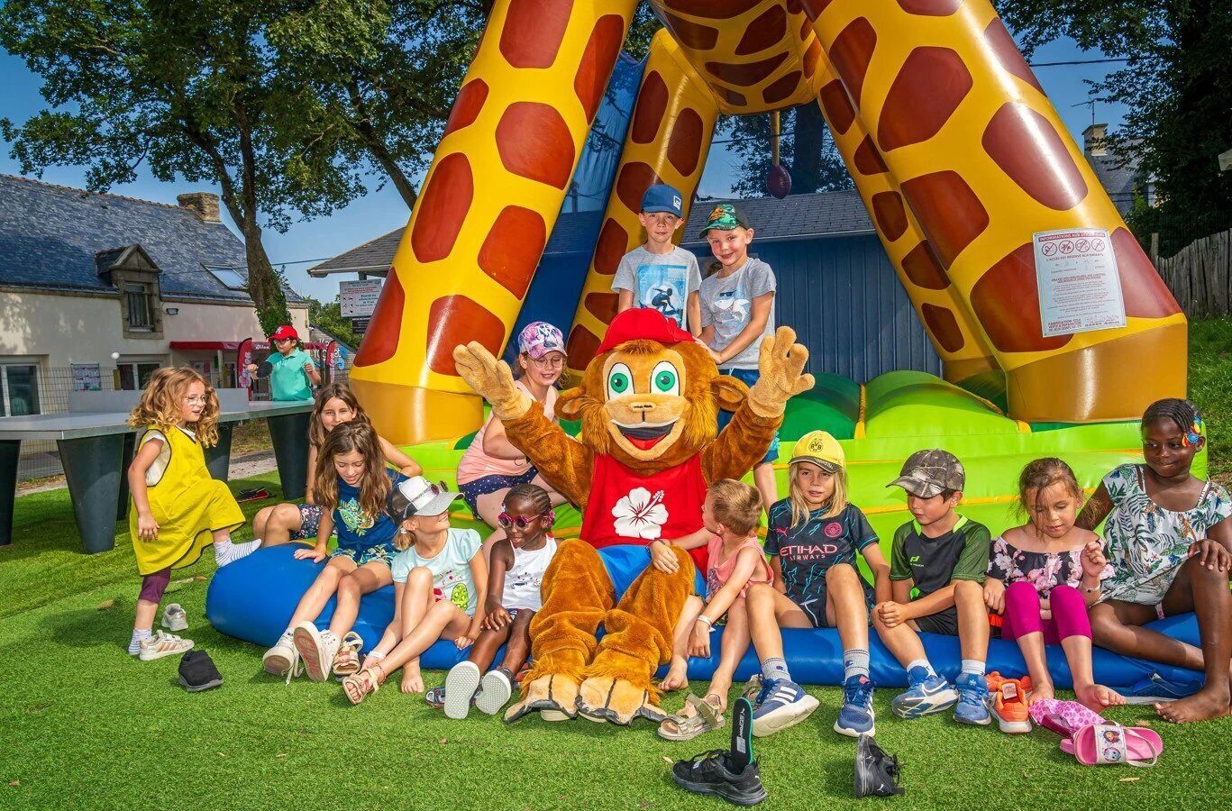 Un grupo de niños y un adulto disfrazado sentados sobre césped, con una gran estructura inflable de jirafa al fondo.