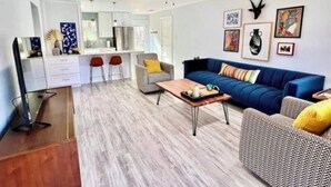 A modern living room with a blue sofa, a grey armchair, and a coffee table. There is a kitchen area with white cabinets and a dining table with red chairs. - Hacienda East - Central + Bright + Just Right (Austin)
