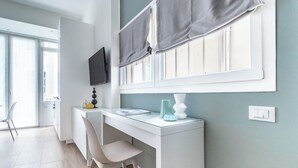 A modern, well-lit room with a white desk, a chair, a flat-screen TV mounted on the wall, and a window with grey curtains. - Pirandello 1 - Studio Apartment, Sleeps 2 (Milano)