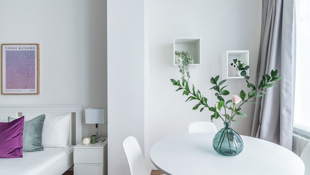 A modern, minimalist hotel room with a neatly made bed, a small bedside table with a lamp, a white round table with a vase of green plants, and a framed artwork on the wall. - Pirandello 1 - Studio Apartment, Sleeps 2 (Milano)