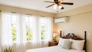 A bedroom with a bed, curtains, a ceiling fan, an air conditioning unit, a nightstand, a lamp, and a potted plant.