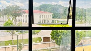 A room with large windows offering a view of a building with arched windows and a red roof, surrounded by greenery. - Flat no Tropical Executive Hotel (Manaus)
