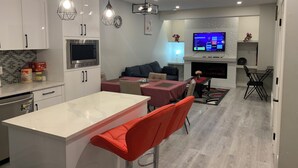 A modern kitchen with white cabinets, a built-in microwave, and a stainless steel refrigerator. There is a dining area with a red table and orange chairs, and a living room with a dark sofa and a flat-screen TV mounted on the wall. - PRIVATE, QUIET, CLEAN LEGAL SUITE FULLY FUNISHED. NEAR DOWNTOWN &TRAIN STATION (Edmonton)