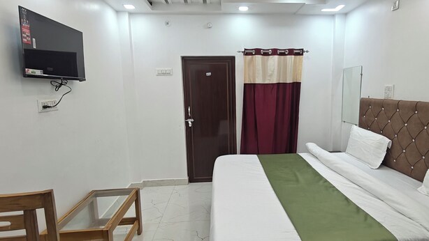 A hotel room with a bed, a TV mounted on the wall, a wooden chair, and a door with a curtain. - Hotel Surya Inn  (Prayagraj)