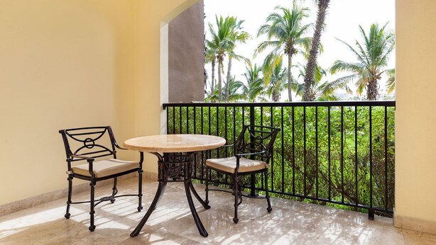 Un balcone con un tavolo rotondo e due sedie, affacciato su un giardino rigoglioso con alte palme.