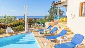 A pool area with blue lounge chairs, a white chair, and a table with a bowl of fruit. - Apollon Dionysus Villa (Peyia)