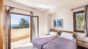 A bedroom with a bed, a nightstand, a window with curtains, and a view of the sea. - Apollon Dionysus Villa (Peyia)