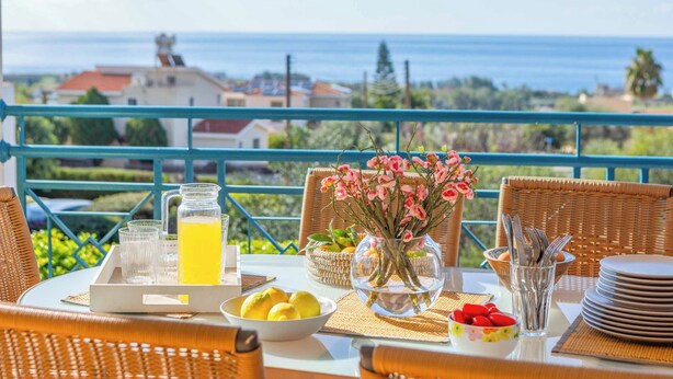 A table set with a pitcher of orange juice, a bowl of lemons, a vase with pink flowers, and a bowl of strawberries on a balcony overlooking the sea. - Apollon Dionysus Villa (Peyia)