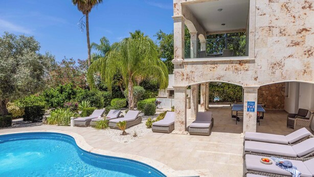 A swimming pool with lounge chairs, a covered patio area, and a stone wall.