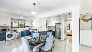 A modern kitchen with white cabinets, a dining area with a glass table, and a view into a bedroom.