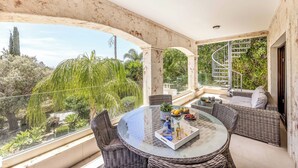 A balcony with wicker furniture, a glass table, and a view of greenery.