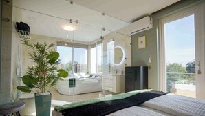 Een moderne slaapkamer met een groot bed, een glazen scheiding, een potplant, een kaptafel met spiegel en uitzicht op de omgeving.