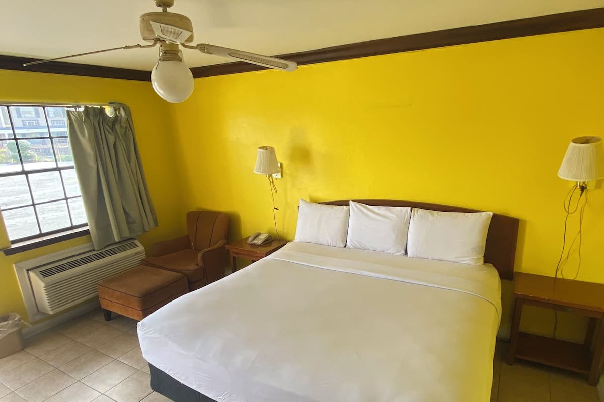 A hotel room with a large bed, a ceiling fan, a window with curtains, a desk with a lamp, and a heater.