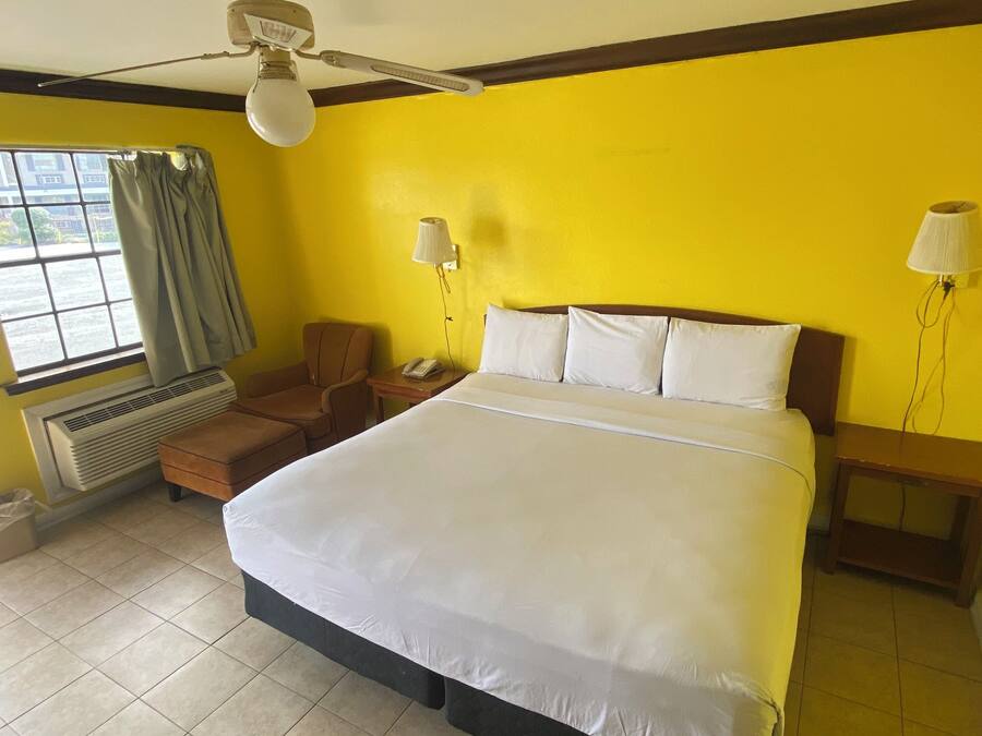 Quarto de hotel com uma cama grande, um ventilador de teto, uma janela com cortinas, uma mesa com luminária e um aquecedor.