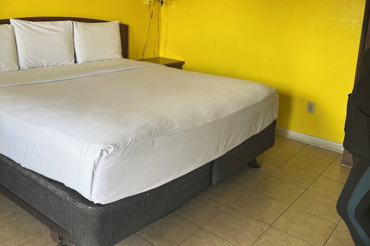 A hotel room with a large bed, a nightstand, a lamp, and a smoke detector on the wall.
