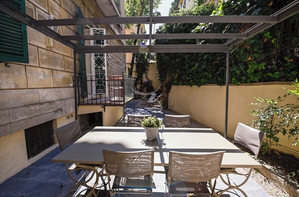 A covered outdoor patio with a table and chairs, a hammock, and a potted plant. - The Muse's Garden (Roma)