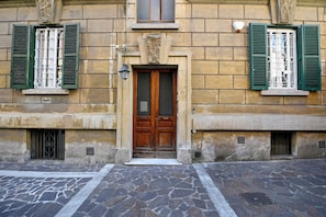 A building with a stone facade, a wooden double door with a glass panel, and green shutters on the windows. - The Muse's Garden (Roma)