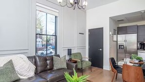 A modern living room with a brown leather sofa, a green armchair, and a dining area with a wooden table and chairs. - LUXE Sun Drenched 2 Bd, Steps to Brown and Wayland (Providence)