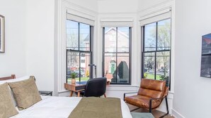 A bedroom with a bed, a desk, a chair, and large windows offering a view of the outside. - LUXE Sun Drenched 2 Bd, Steps to Brown and Wayland (Providence)
