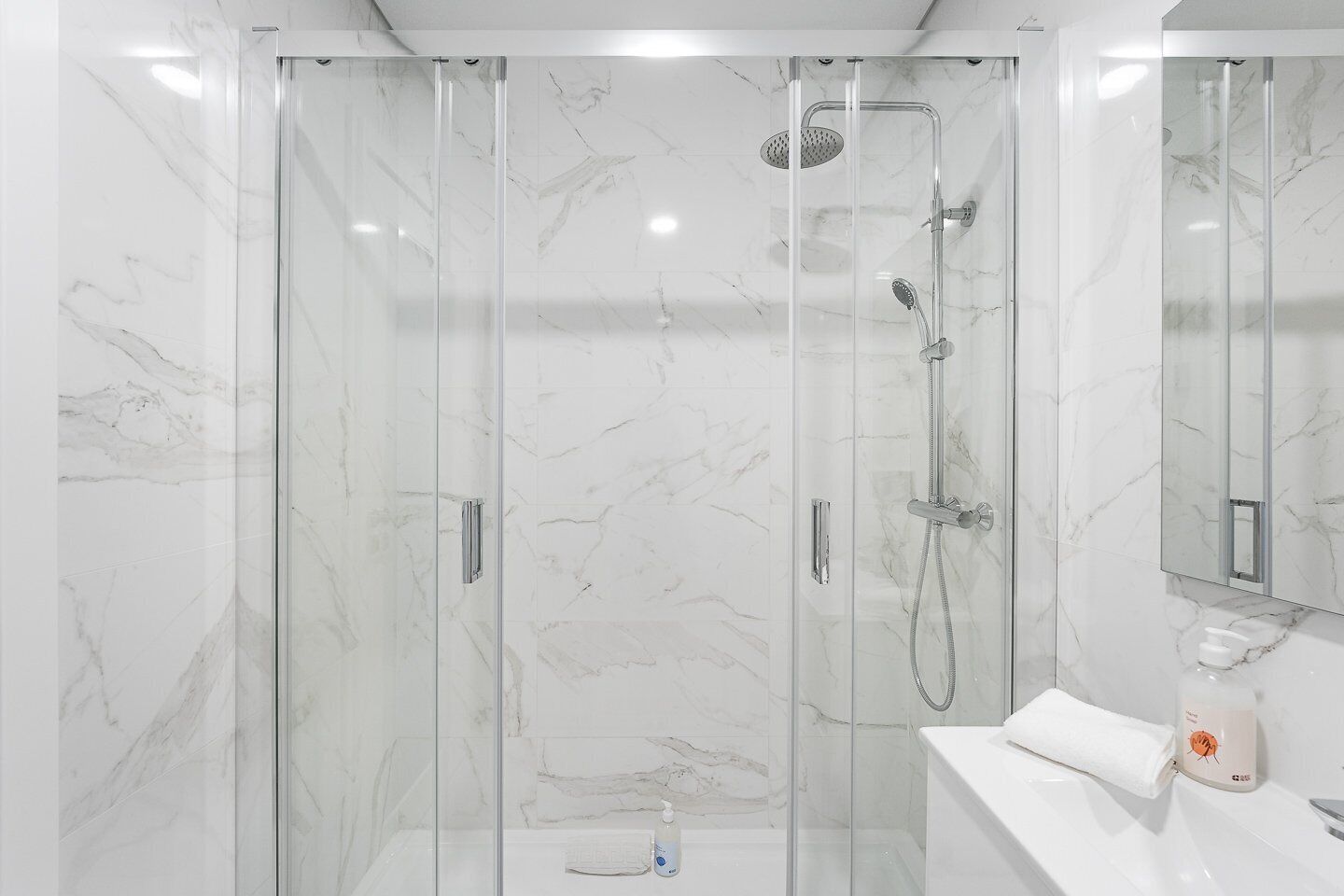 Un baño moderno con ducha de vidrio, pared de mármol blanco y lavamanos blanco con una toalla.
