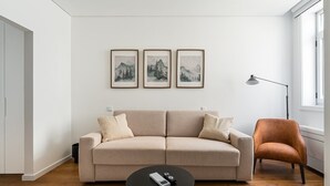 A modern living room with a beige sofa, a small black coffee table, and a brown armchair. There are three framed pictures on the wall above the sofa. - GuestReady - Inviting Hideaway in Matosinhos (Matosinhos)