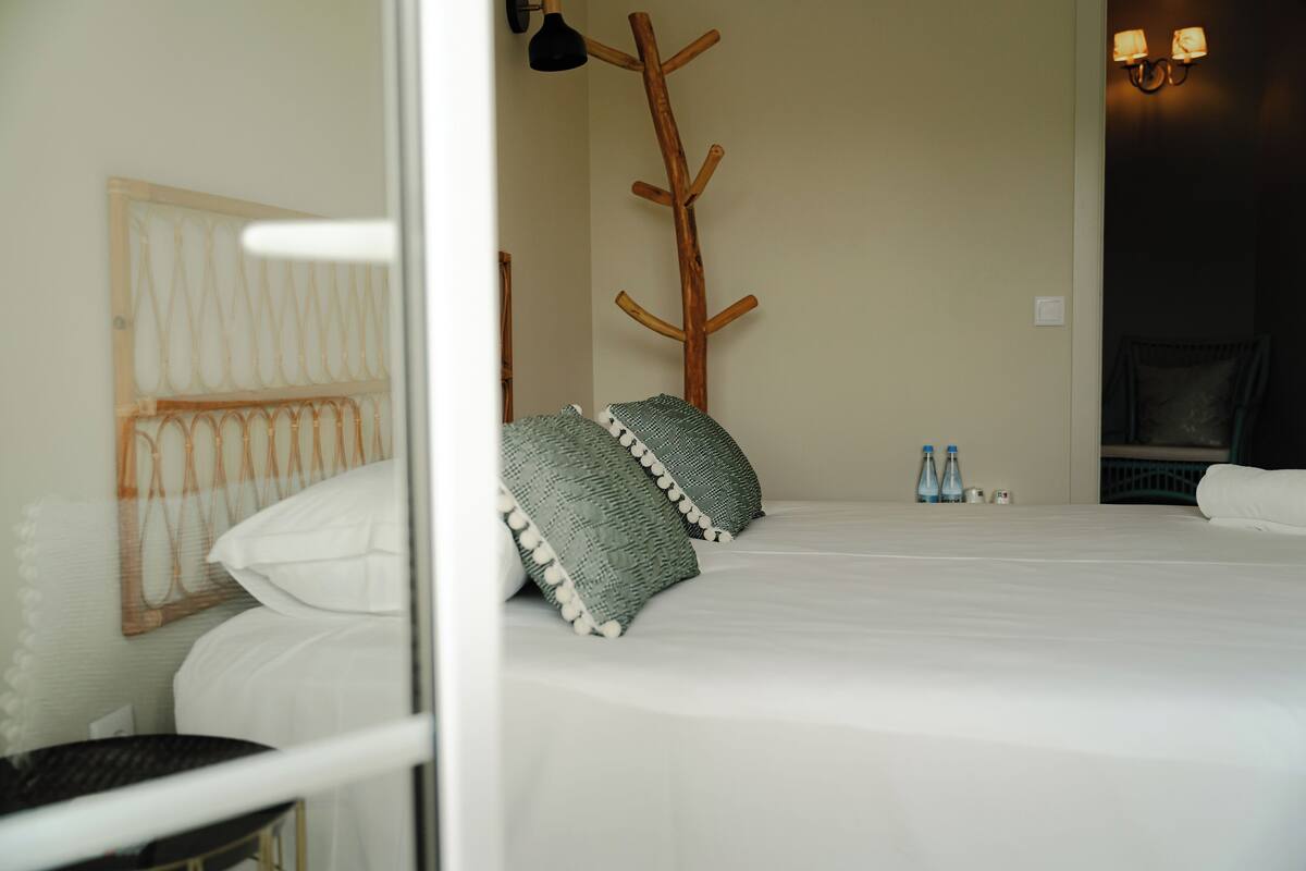 Un lit bien fait, avec du linge de lit blanc et deux coussins verts, un porte-manteau en bois et une lampe fixée au mur.