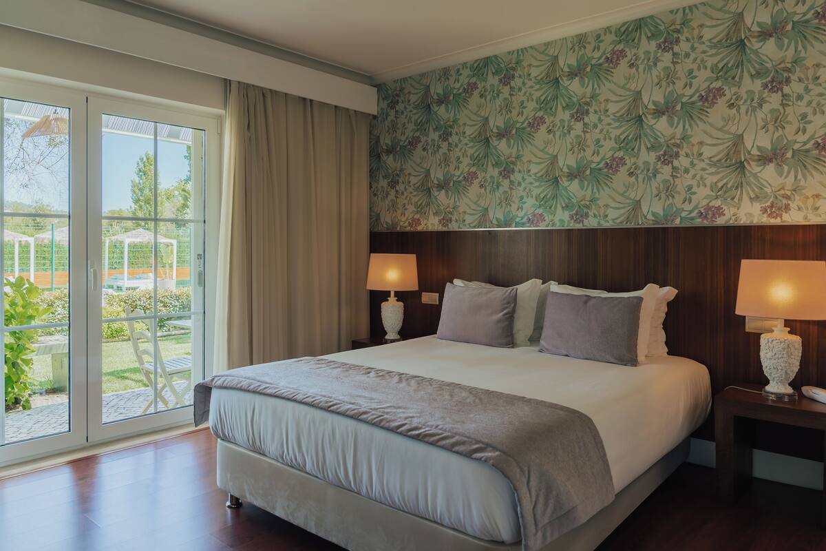 Une chambre d’hôtel avec un grand lit, deux tables de chevet avec des lampes, un papier peint à motifs floraux et une porte-fenêtre coulissante donnant sur l’extérieur.
