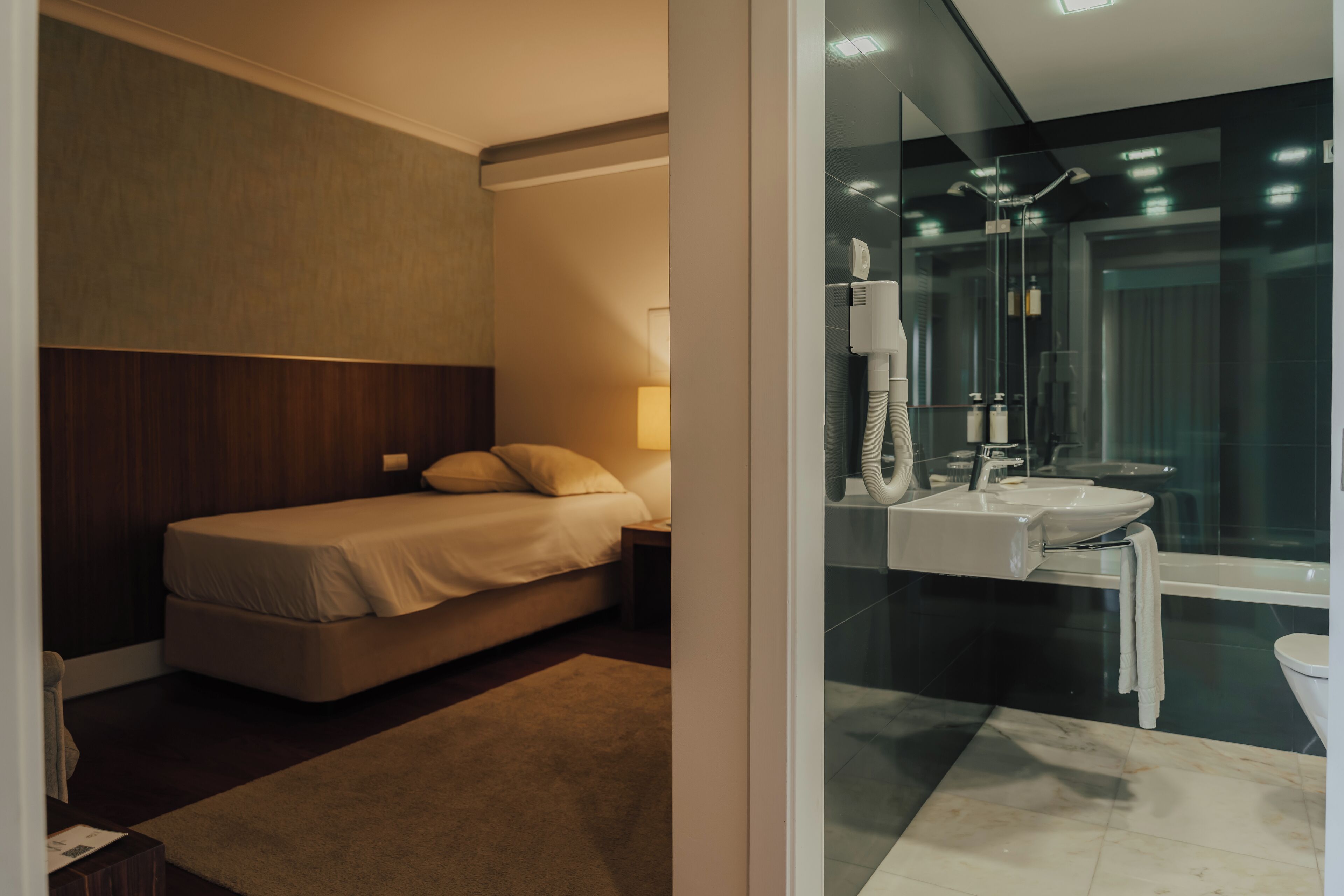 A hotel room with a bed, bedside table, and a bathroom with a sink, mirror, and toilet.