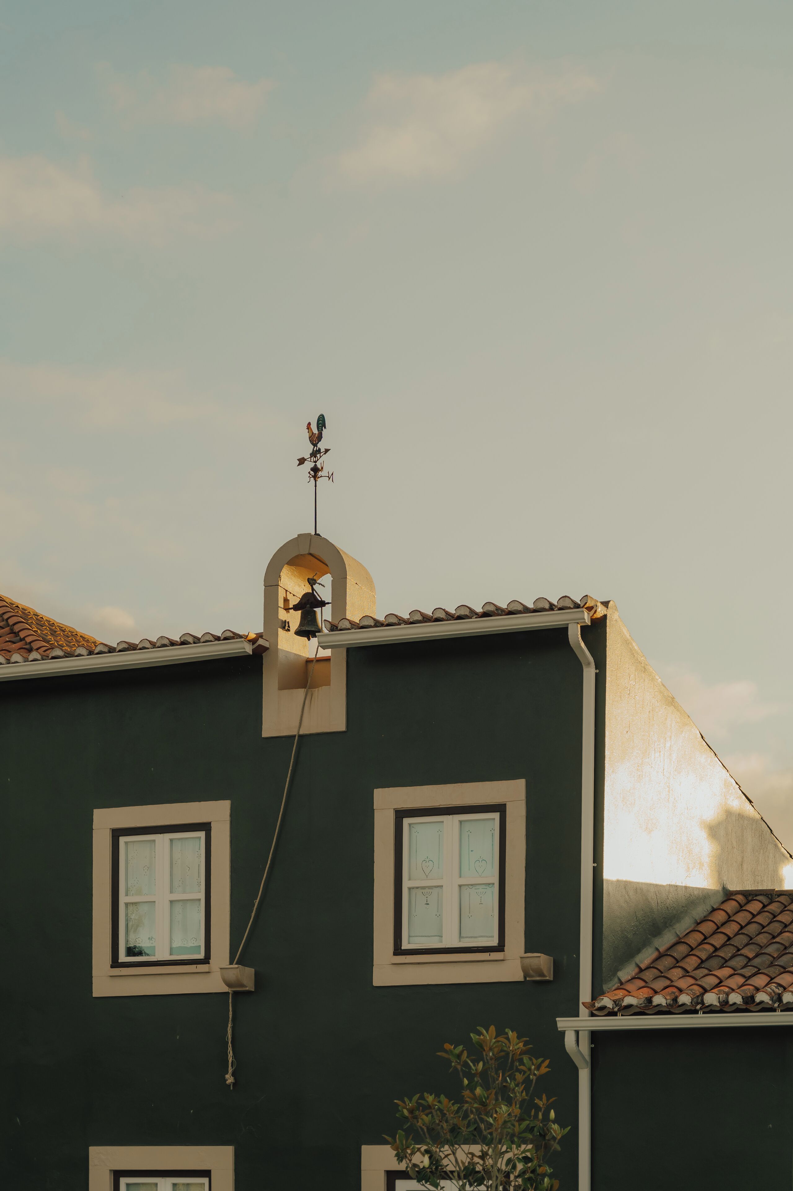 A building with a weather vane on the roof, a small window with a decorative arch, and a potted plant on the ground.