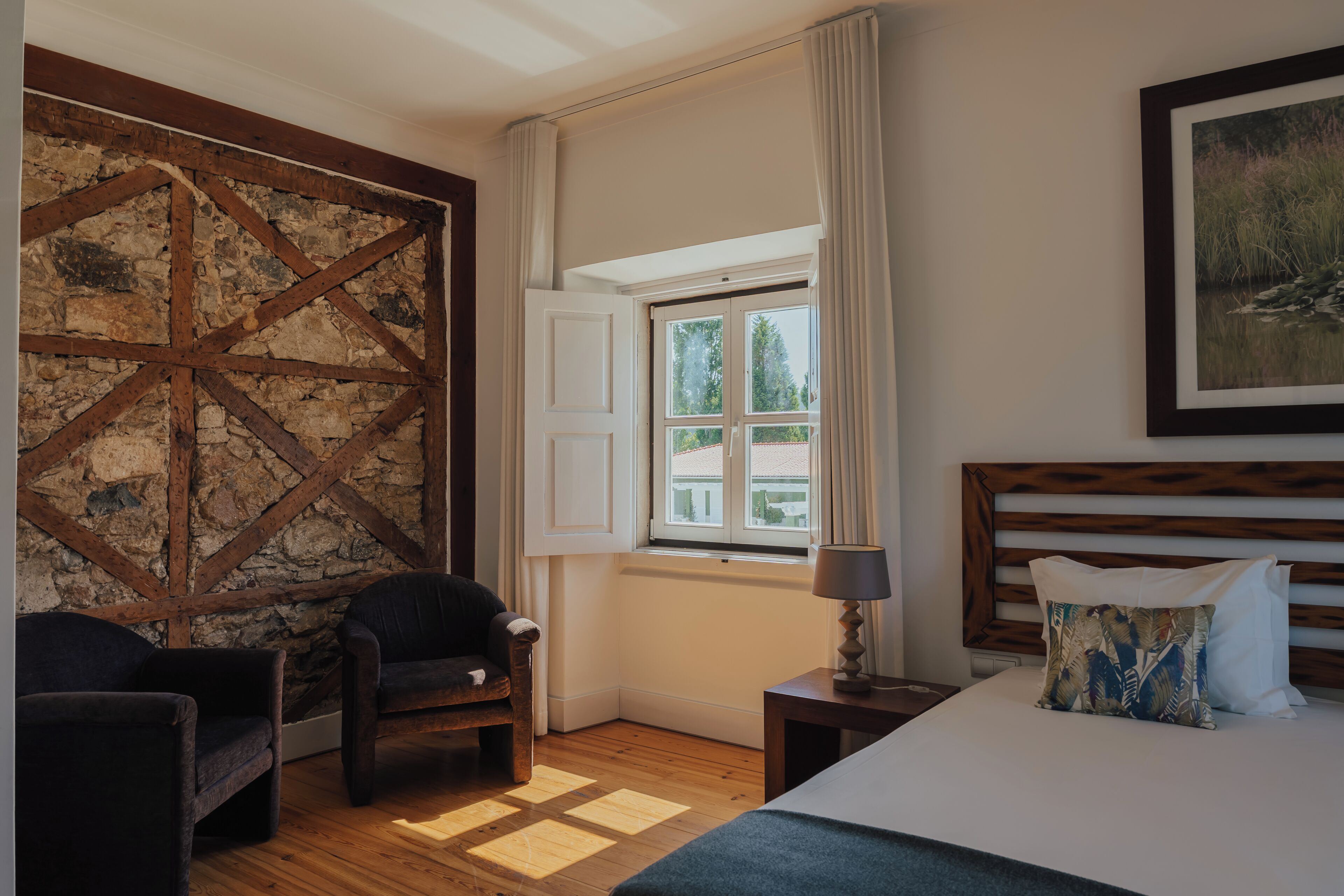 Een kamer met een bed, een stoel, een nachtkastje, een raam met gordijnen en een muur met een houten balkenpatroon.
