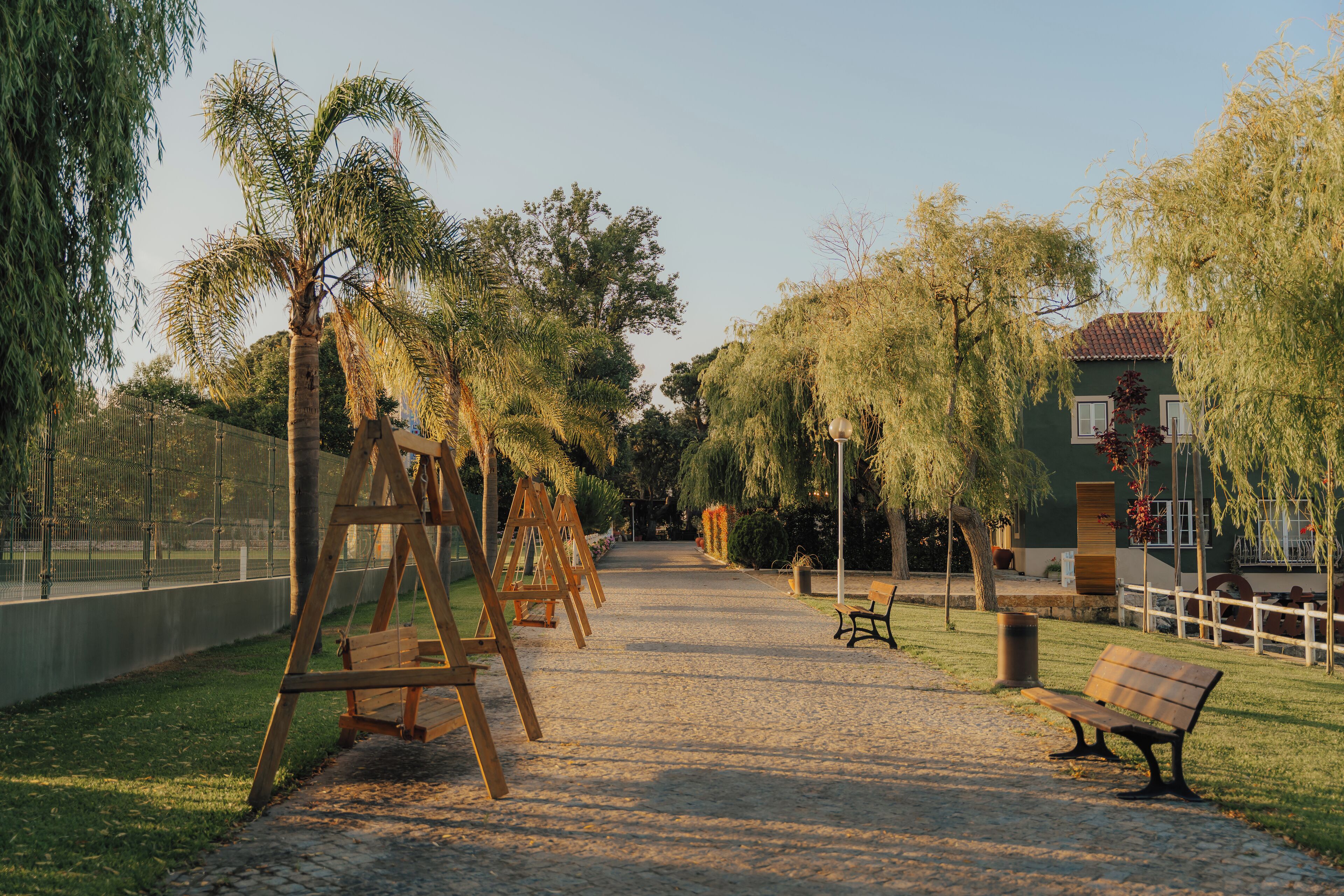 Un sendero en un parque con columpios y bancos, rodeado de árboles y una valla.