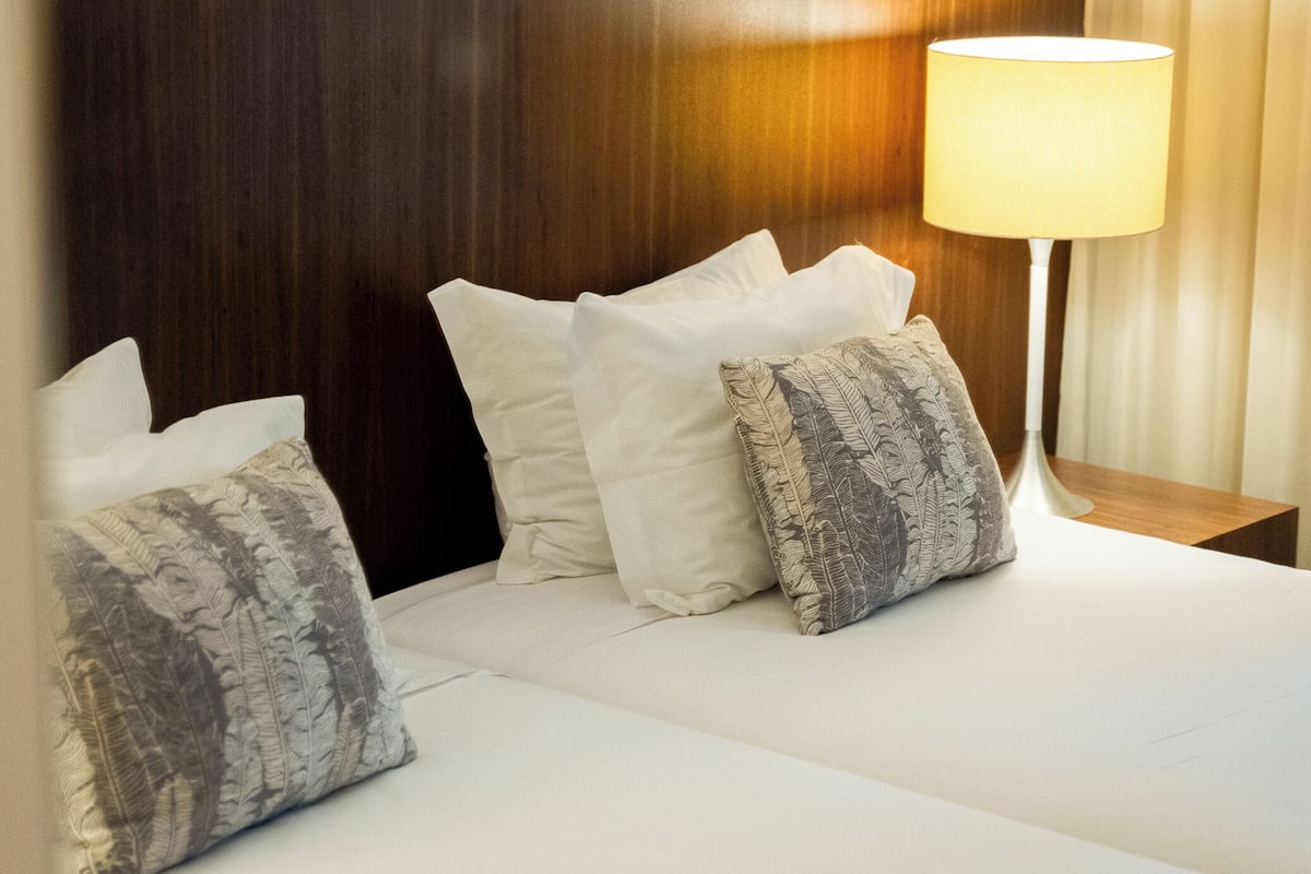 Un lit bien fait, avec du linge de lit blanc et des coussins décoratifs, une tête de lit en bois et une lampe de chevet.