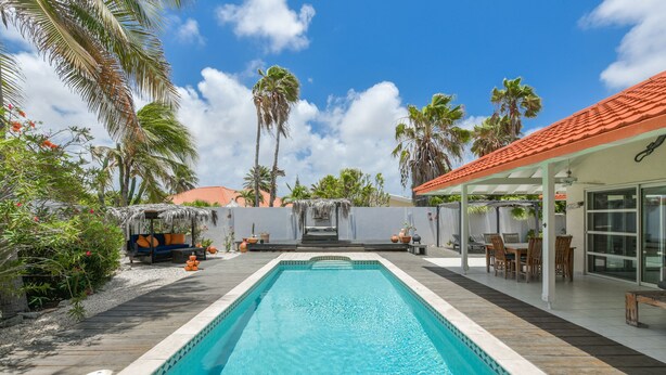 A swimming pool with a wooden deck, a covered seating area, and palm trees in the background.