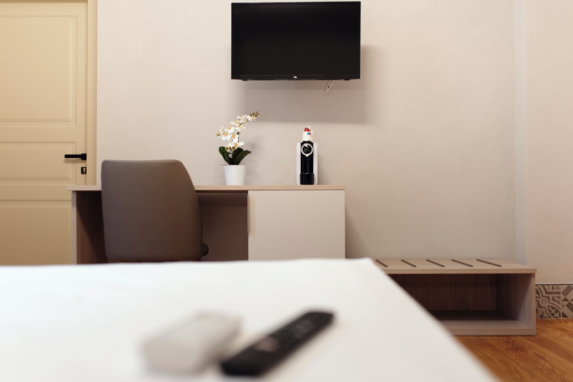 Una habitación de hotel moderna con una televisión de pantalla plana montada en la pared, un pequeño escritorio con una planta en maceta, un control remoto sobre la cama y suelo de madera.