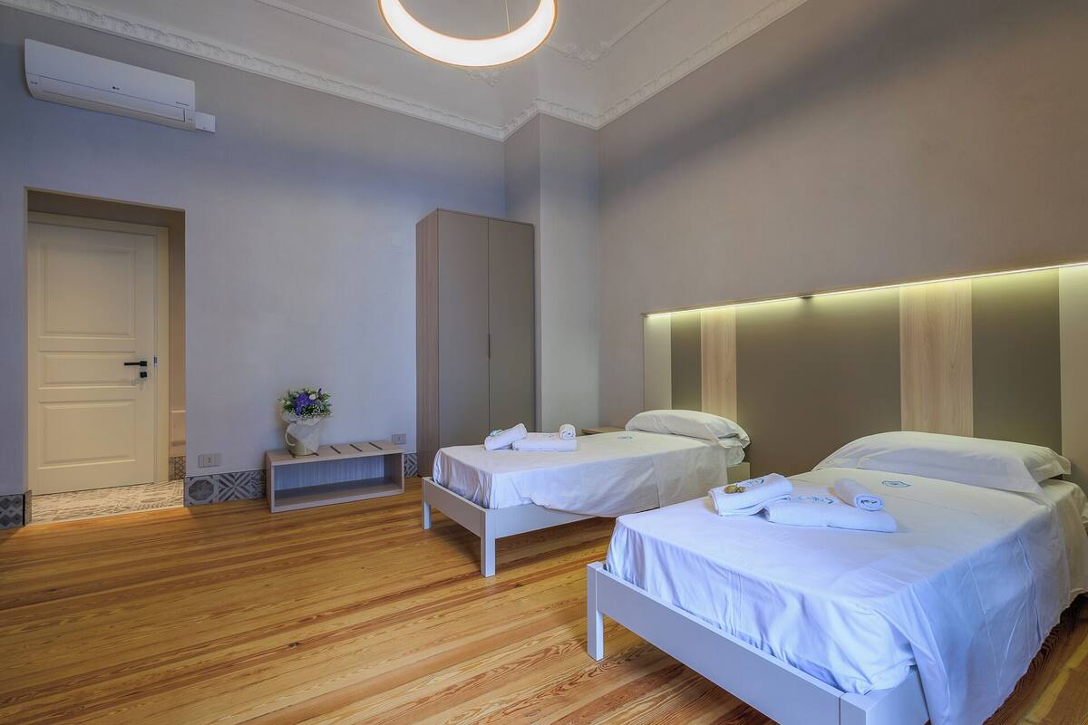 A hotel room with two beds, a wooden floor, a white door, and a wall-mounted air conditioner.