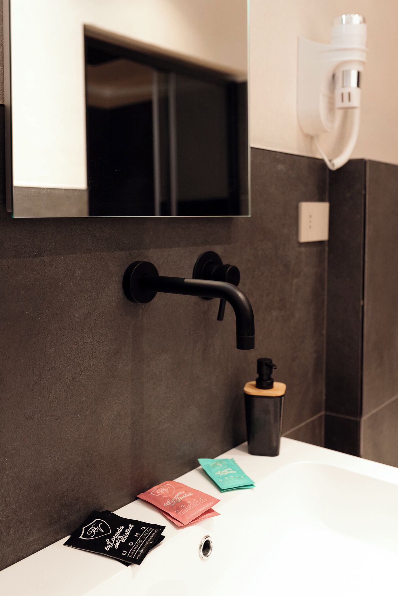 Un bagno moderno con un rubinetto nero, uno specchio e un lavandino bianco. Sul lavandino ci sono tre piccole borse.