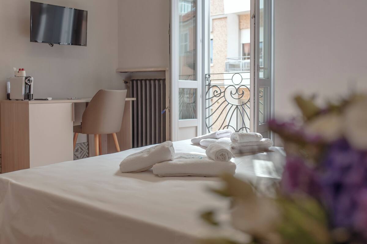 A hotel room with a bed, a desk, a chair, a TV, and a balcony with a railing.